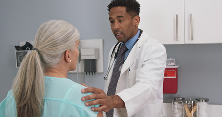 Millennial black doctor comforting senior patient by placing his hand on her shoulder. Elderly white woman receiving support from young male doctor 