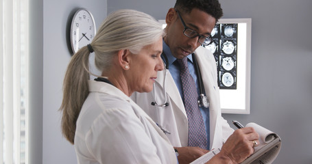 Senior female doctor taking notes of patients ct scan while consulting with young colleague. Elderly medical physician writing on clipboard and talking to young assistant