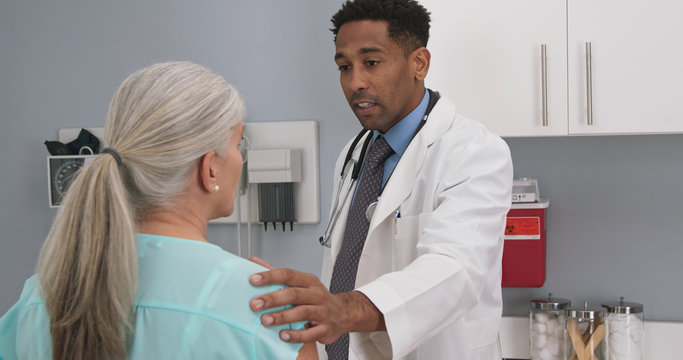 Millennial Black Doctor Comforting Senior Patient By Placing His Hand On Her Shoulder. Elderly White Woman Receiving Support From Young Male Doctor 