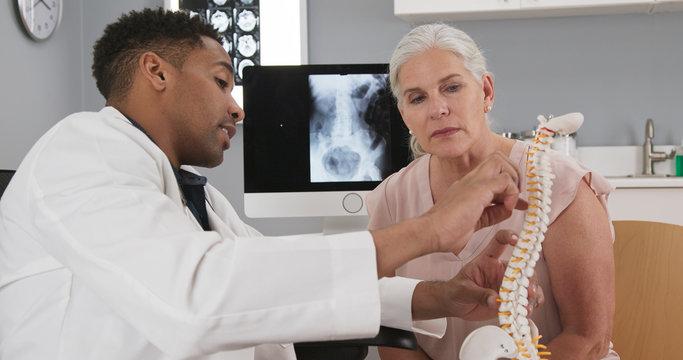 Senior female patient consulting with young doctor about back injury. Close up of black doctor holding model of spinal cord and explaining to elderly woman source of her pain - Powered by Adobe