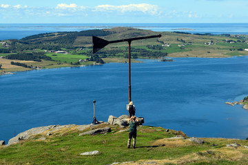 Two people stand on top of a green hill. A man and a woman look at the sea and the settlement on the shore. View from the back. Tourism as a way of life. Life and travel in Norway.