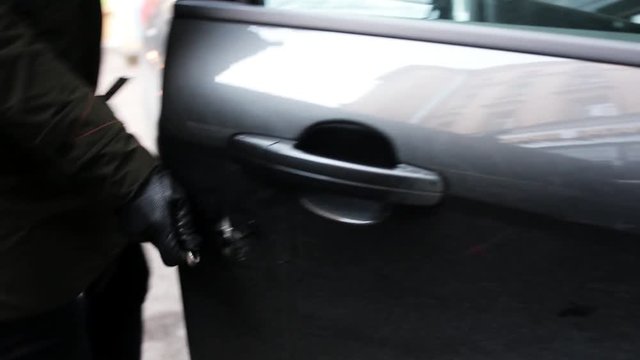 Woman wearing black leather gloves and holding thin metallic stick with curved tip is aggressively making big hole in surface of car door at street in day light and opens it.