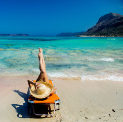 Young redhead girl in black bikini and with hat lying down on lounger on Balos beach, west Crete, Greece. Summertime season vacation, July