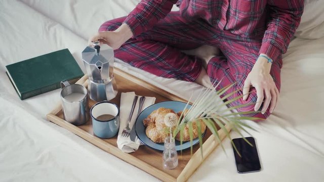 Female having breakfast in bed Preparing or pouring and drinking milk and hot coffee food in tray or morning meal in bedroom wearing checkered pajamas after wake up and check smartphone app network