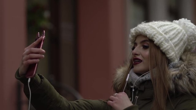 Cute girl in knitted hat and parka with fur is talking and smiling for Instagram stories. Girl is holding her phone in front of her and wearing an earphones.