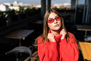 Lifestyle portrait of smiling cute woman in red knitted sweater and tylish sunglasses at Outdoor terrace. Sunny fall season. Romantic mood.