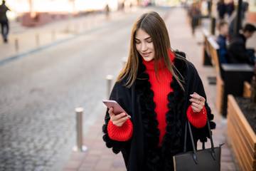 Portrait of young woman in coat and hat using smart phone while standing on city street.
