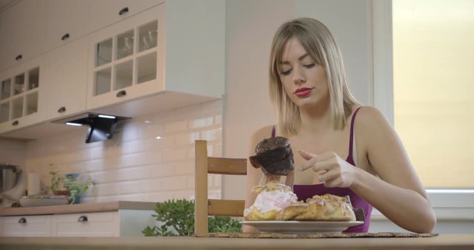 Medium shot of a young attractive blond woman sitting at her kitchen table with a plate full of desserts stuffing her face with sweets