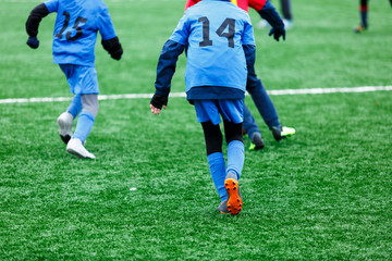 Obraz premium Boys in red and blue sportswear plays soccer on green grass field. Youth football game. Children sport competition, kids plays outdoor, winter activities, training