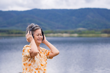 Portrait of happy senior woman listening music on headphone for relaxing with side the lake. Relaxing time concept.