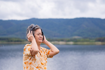 Portrait of happy senior woman listening music on headphone for relaxing with side the lake. Relaxing time concept.
