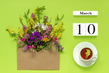 Wooden cubes calendar March 10. Cup of herbs tea, kraft envelope with multi colored flowers on...