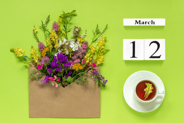 Wooden cubes calendar March 12. Cup of herbs tea, kraft envelope with multi colored flowers on green background. Concept hello spring Creative Top view Flat lay
