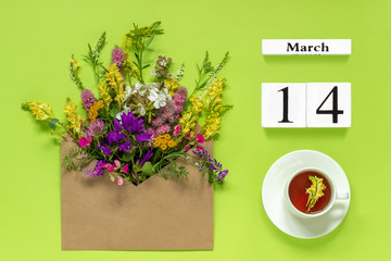 Wooden cubes calendar March 14. Cup of herbs tea, kraft envelope with multi colored flowers on green background. Concept hello spring Creative Top view Flat lay