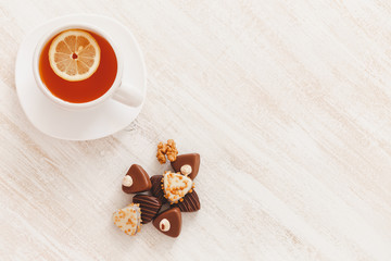 Chocolate candy with nuts, cup of hot tea with lemon and saucer on white wooden table. Flat lay, top view.