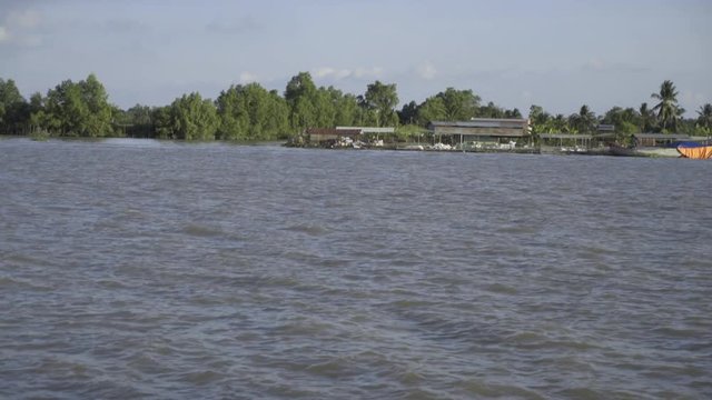 Pan up to a fish farm and boats on the shore of a river in Southeast Asia on a beautiful partly cloudy day; view from a passing boat.