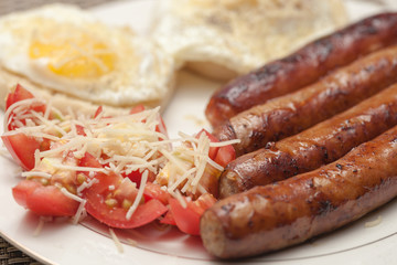Fried rustic sausage links with sunny side up eggs with tomato and shreded aged cheese salad