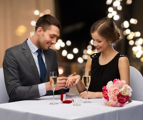 proposal, valentines day and people concept - happy young man giving woman engagement ring at restaurant over festive lights on background