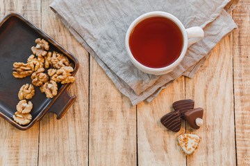 Fototapeta na wymiar Cup of hot black tea with chocolate candy and walnuts on wooden table. Flat lay, top view.
