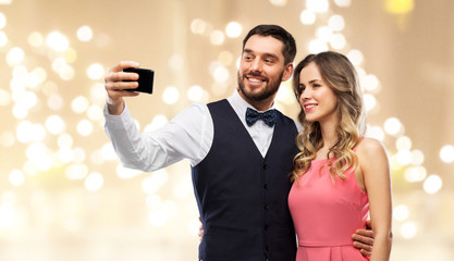 technology and people concept - happy couple in party clothes taking selfie by smartphone over beige background with festive lights