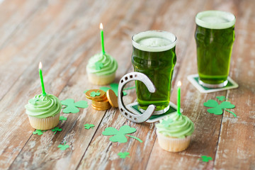 st patricks day, holidays and celebration concept - glass of green beer, cupcakes with candles, horseshoe and gold coins on wooden table