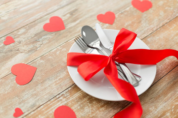 valentines day, table setting and festive dinner concept - plate with spoon, knife and fork tied with red ribbon on wooden background decorated by paper hearts