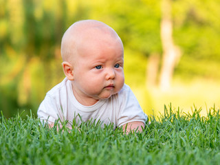 The baby lies or crawls along the green lawn at sunset and is looking ahead seriously. Emotions baby. Vacation with children. Childcare. Goods for kids. Sun protection. Child development.