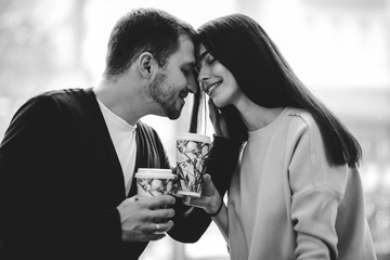 Loving couple is siting face to face on the background of window and holding cups in their hands. Black and white photo