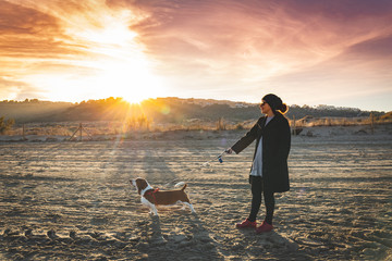 Girl walking with her dog with beautiful winter sunset. Woman walking with her Basset Hound in a coat and cap on the beach