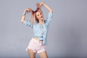 Beautiful young woman expresses happy emotions, has broad pleasant smile, glad to new unusual pink color of his hair. Smiling overjoyed female in blue denim shirt enjoys over gray background