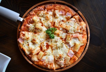 Pizza topped with tomato sauce, cheese and pineapple serve on wooden plate on wooden table. Photo of Hawaiian pizza.