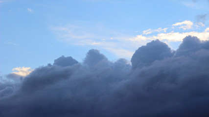 Textured gray clouds on the blue sky. In the background are white clouds illuminated by the setting sun. Summer evening.