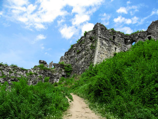 Fototapeta na wymiar Stone ruined walls of Khust fortress are overgrown with grass - bottom view. The gloomy gray remnants of a medieval castle against the backdrop of bright spring greenery and blue sky