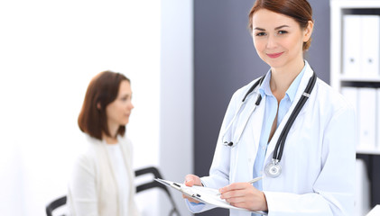 Fototapeta premium Doctor woman at work. Portrait of female physician filling up medical form while standing near reception desk at clinic or emergency hospital. Patient woman sitting at the background. Medicine concept