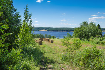Fototapeta na wymiar Beautiful summer landscape with sloping meadow, hay, forest and river with blue sky with clouds.