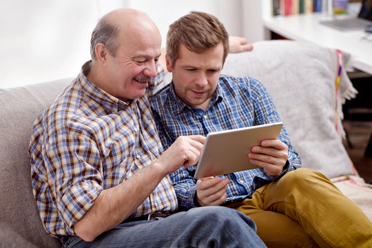 Pleasant Senior Man Pointing At The Tablet, Showing An Interesting Article To His Son