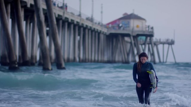 Surfer Smiles And Walks Toward Camera After Surf Session, Next To Pier At Sunset (Slow Motion)