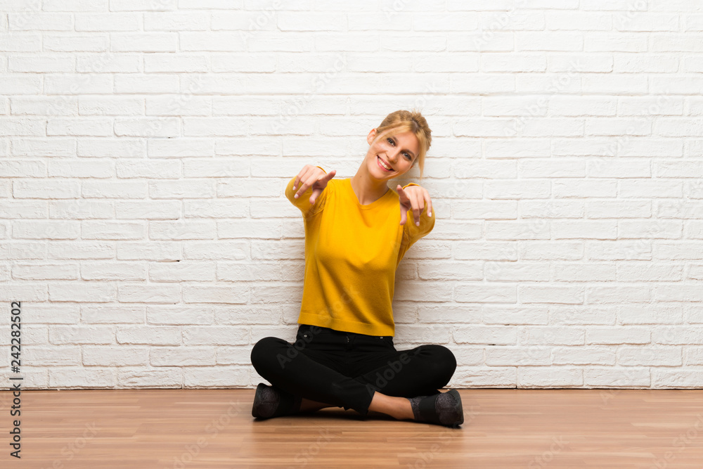 Wall mural young girl sitting on the floor points finger at you while smiling