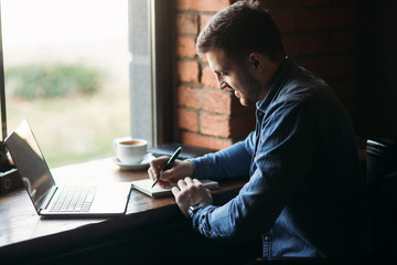 Man with a beard uses a laptop in a cafe and writes a note