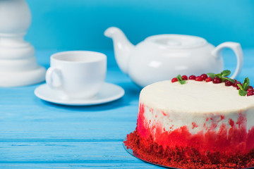 cake decorated with red currants and mint leaves between white cup, tea pot and stand isolated on blue