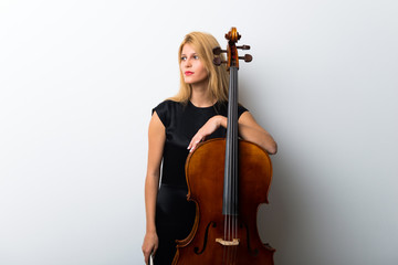 Young blonde girl with her cello posing on white wall