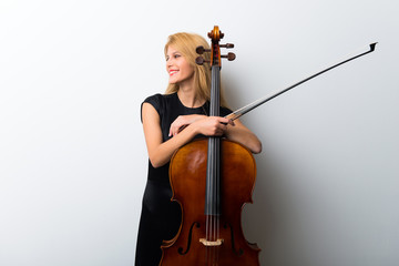 Young blonde girl with her cello posing on white wall
