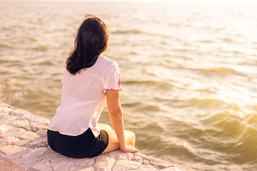 Fototapeta na wymiar Young woman sitting on stone wall enjoying herself watching sunset at the beach