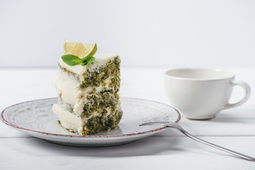 piece of white cake decorated with lime slice and mint leave on saucer near cup isolated on white