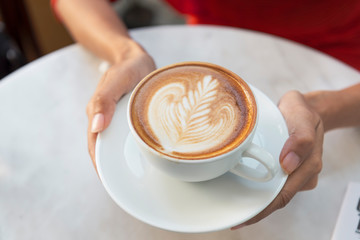 Hand holding latte or Cappuccino with frothy foam, coffee cup top view on table in cafe.