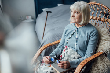 pensive senior woman sitting in wicker rocking chair with book at home