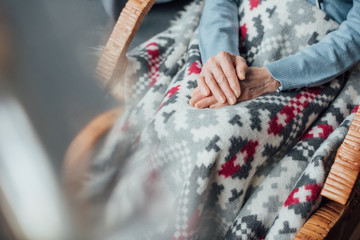 cropped view of senior woman sitting in wicker rocking chair with blanket and folded hands at home
