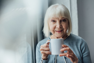 smiling senior woman holding cup of coffee at home and looking at camera