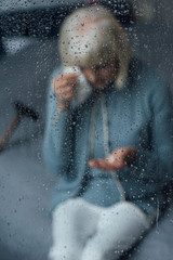 Fototapeta na wymiar senior woman sitting on bed and wiping tears with handkerchief at home through window with raindrops