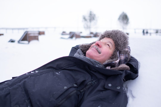 Senior Handsome Man In Warm Clothes And Hat Lying On Snow And Dreaming